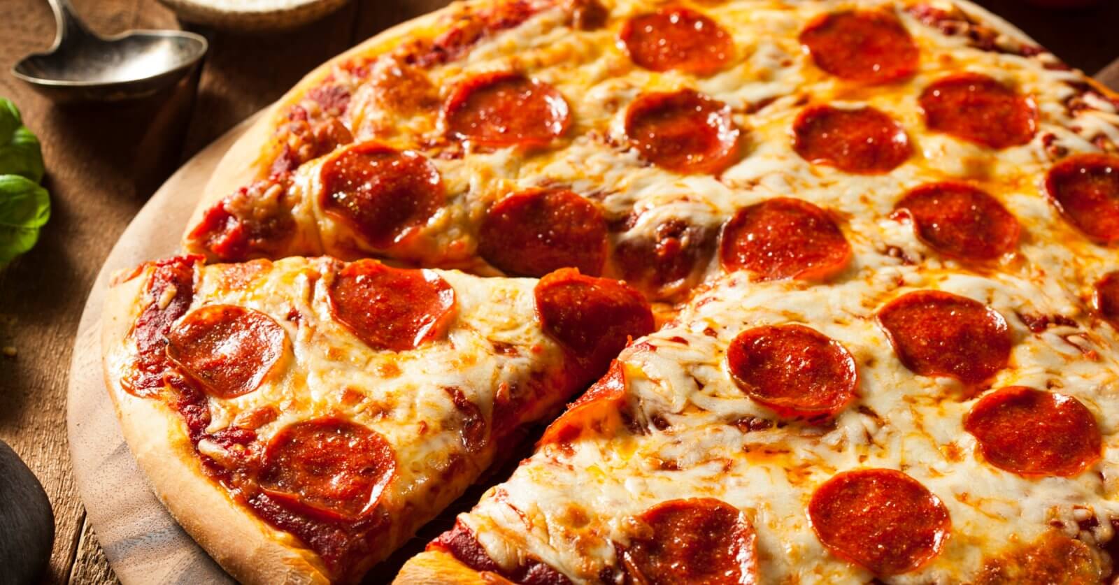 A close-up of a pepperoni pizza on a wooden surface. The pizza has a golden-brown crust and is topped with melted cheese and evenly distributed pepperoni slices. One slice is partially separated from the rest, showcasing the gooey cheese and crisp edges. Leafy greens and utensils are blurred in the background, reminiscent of a Sulit Dining Experience at SM Manila's best restaurants.