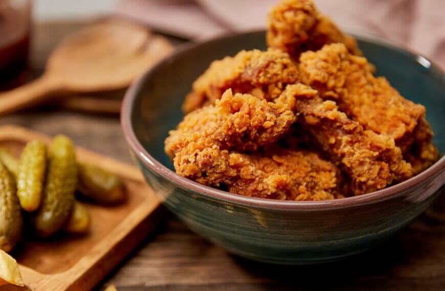 A bowl filled with crispy fried chicken pieces sits on a wooden surface. Beside it, a wooden tray holds several pickles and some fries. In the background, a wooden spoon and a jar of dipping sauce can be seen. This cozy, rustic scene captures the essence of comfort food you'd find at one of the best restaurants in SM Podium Mall.
