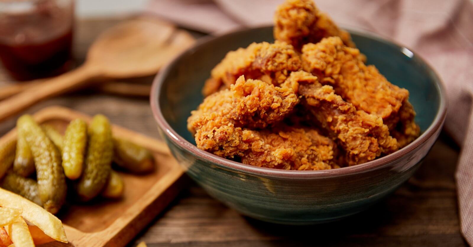 A bowl filled with crispy fried chicken pieces sits on a wooden surface. Beside it, a wooden tray holds several pickles and some fries. In the background, a wooden spoon and a jar of dipping sauce can be seen. This cozy, rustic scene captures the essence of comfort food you'd find at one of the best restaurants in SM Podium Mall.
