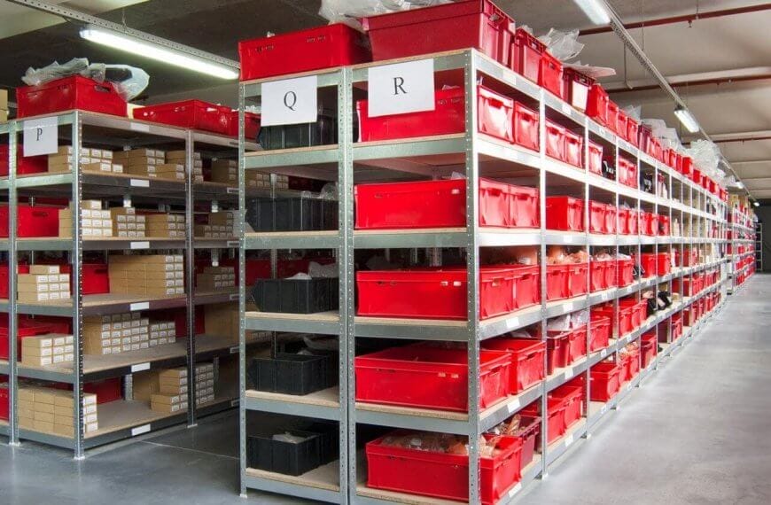 A well-organized warehouse aisle in Metro Manila features metal shelving units filled with red and black plastic storage bins. Each shelf has white labels with letters "Q" and "R" indicating storage categories. Companies rely on these efficient storage solutions, where cardboard boxes and items are neatly arranged in the bins. The floor is clean and polished.