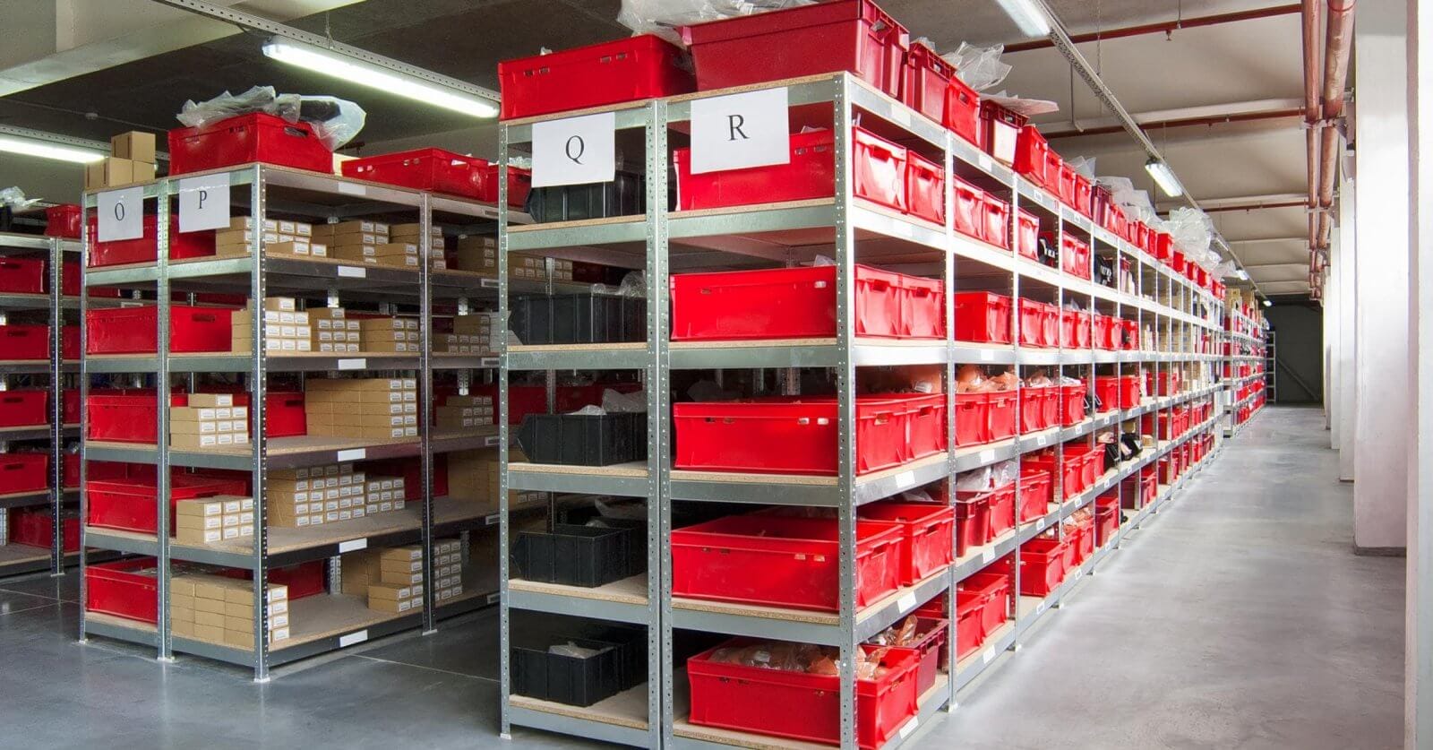 A well-organized warehouse aisle in Metro Manila features metal shelving units filled with red and black plastic storage bins. Each shelf has white labels with letters "Q" and "R" indicating storage categories. Companies rely on these efficient storage solutions, where cardboard boxes and items are neatly arranged in the bins. The floor is clean and polished.