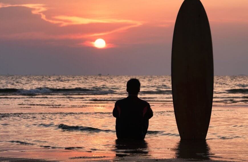 A person, seen from behind, sits on wet sand at the beach during sunset. Next to them, a surfboard is positioned upright in the sand. The sky is filled with vibrant hues of orange and pink as the sun sets over the calm ocean waves in the background, evoking memories of days at surfing school.