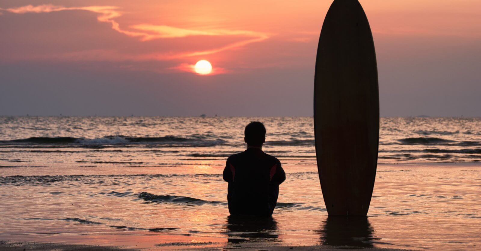A person, seen from behind, sits on wet sand at the beach during sunset. Next to them, a surfboard is positioned upright in the sand. The sky is filled with vibrant hues of orange and pink as the sun sets over the calm ocean waves in the background, evoking memories of days at surfing school.