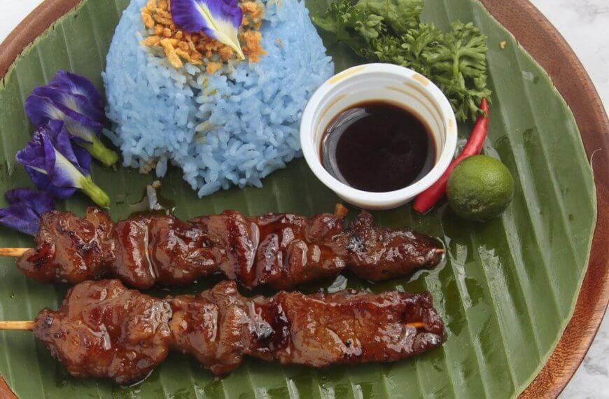 A plate with two bbq and blue rice garnished with crispy bits and an edible flower, a small cup of dark dipping sauce, a lime, a chili, and parsley served on a large banana leaf. Celebrated among the 50 Best Restaurants near UP Diliman, this dish is presented on a wooden tray against a light background.
