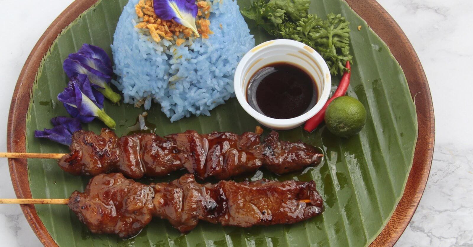 A plate with two bbq and blue rice garnished with crispy bits and an edible flower, a small cup of dark dipping sauce, a lime, a chili, and parsley served on a large banana leaf. Celebrated among the 50 Best Restaurants near UP Diliman, this dish is presented on a wooden tray against a light background.
