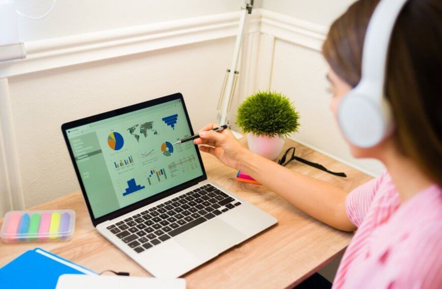 Virtual Assistant wearing headphones sits at a desk in Cebu, focusing on a laptop displaying various graphs and charts. They are pointing to the screen with a pen. The workspace includes colorful sticky notes, a pot of pens, and a small potted plant. The room is well-lit from a nearby window, perfect for productivity with the best virtual assistants.