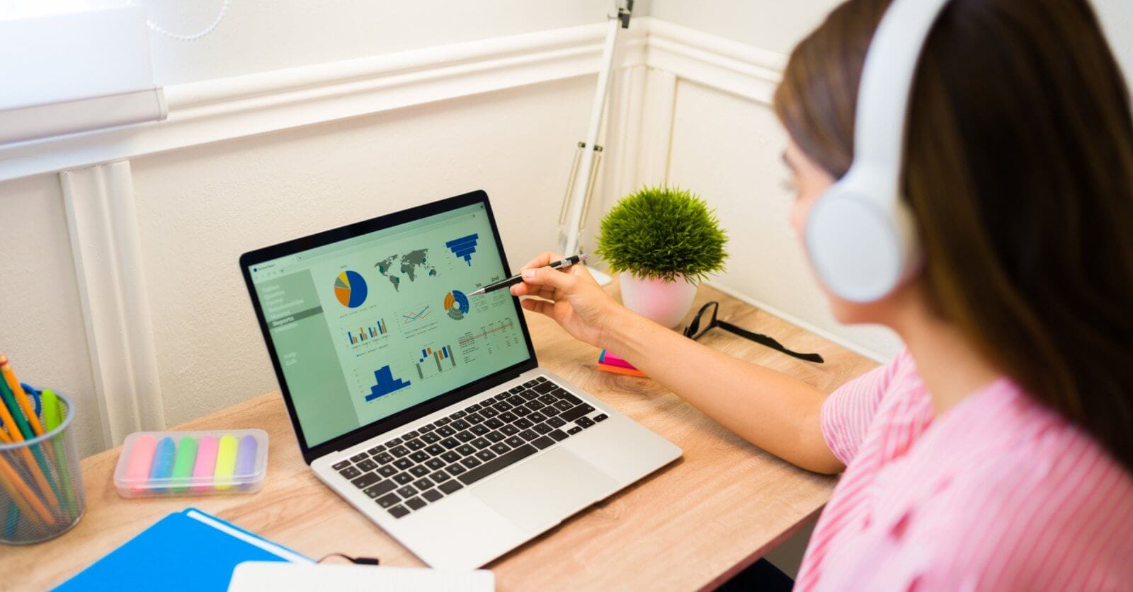 Virtual Assistant wearing headphones sits at a desk in Cebu, focusing on a laptop displaying various graphs and charts. They are pointing to the screen with a pen. The workspace includes colorful sticky notes, a pot of pens, and a small potted plant. The room is well-lit from a nearby window, perfect for productivity with the best virtual assistants.