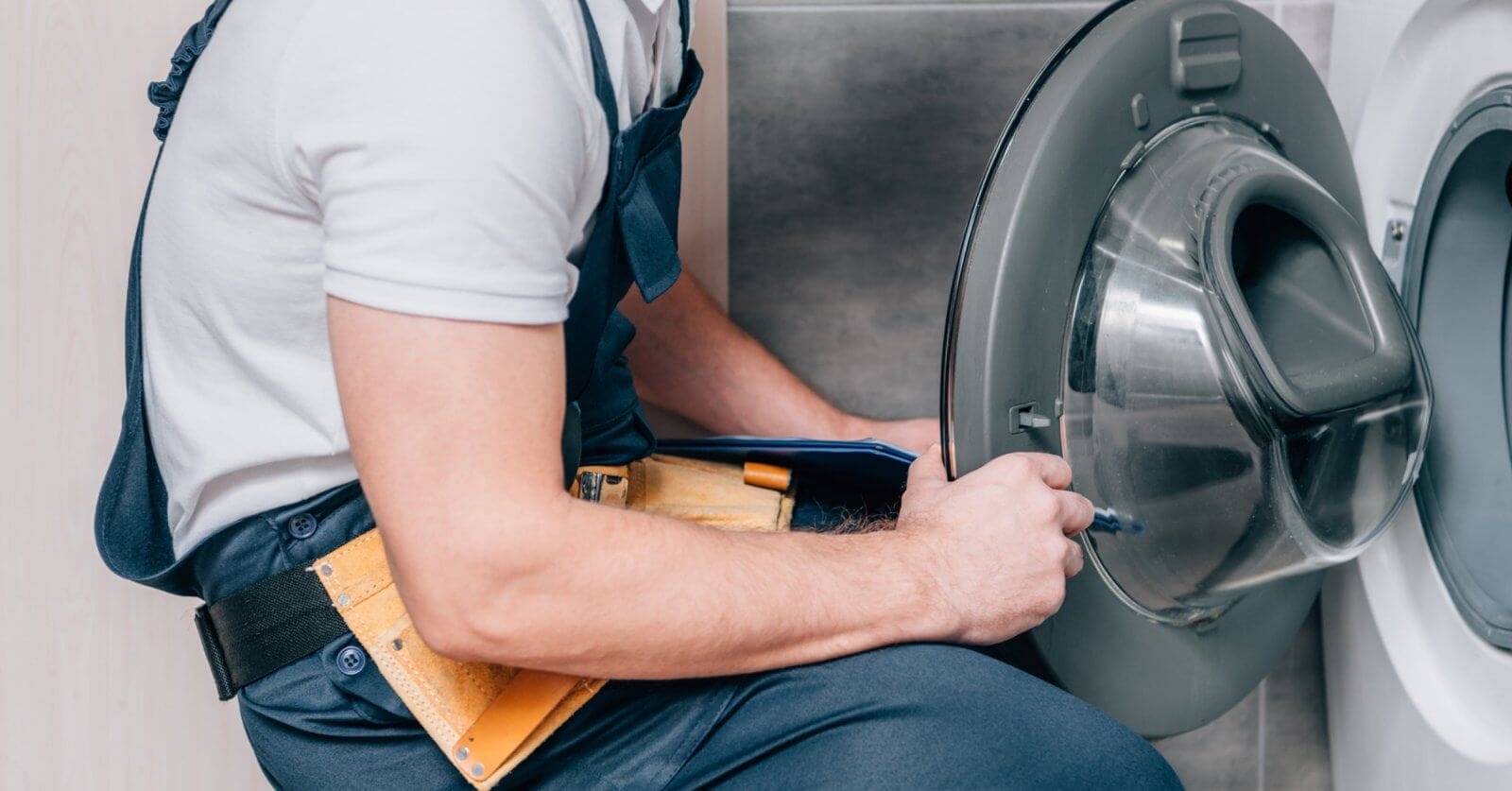 A person repairing a front-loading washing machine in Metro Manila. With a yellow tool belt brimming with various tools around their waist, they are closely examining the open door of the washing machine, appearing focused on providing top-notch Washing Machine Repair Services.