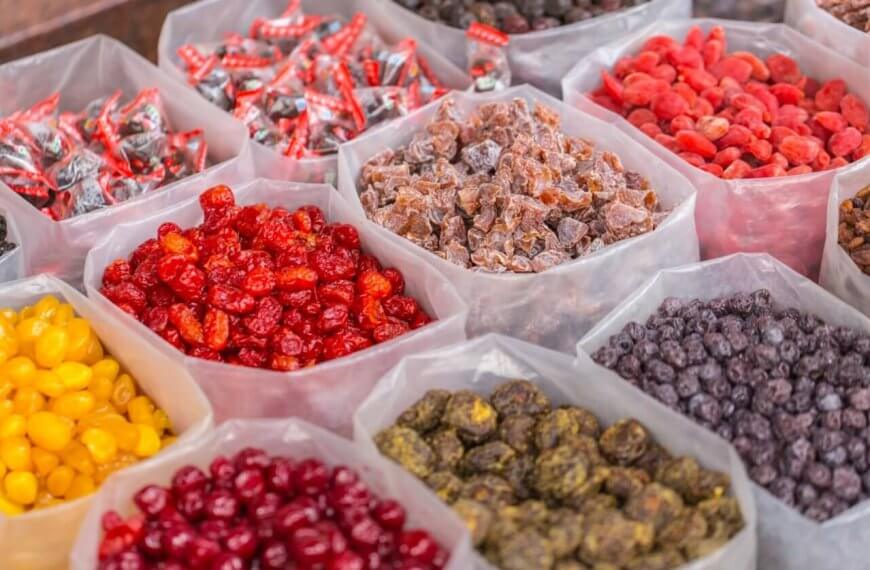 A market display of various dried fruits and candies from the best wholesale food suppliers in Metro Manila. Clear plastic bins are filled with colorful selections such as red cherries, yellow berries, purple raisins, and multicolored preserved fruits. Wrapped candies are also visible in the background, creating a vibrant and inviting scene.