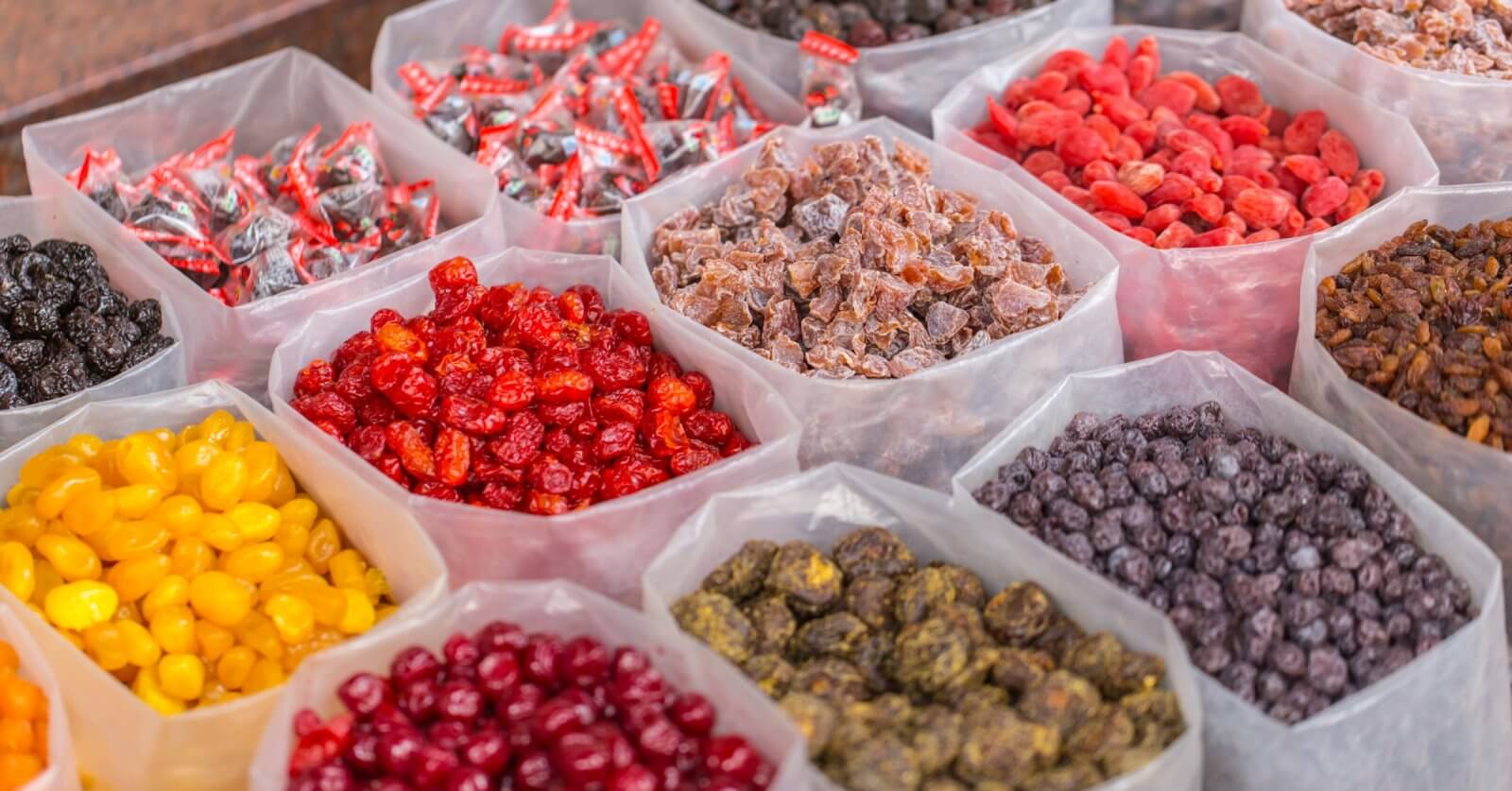 A market display of various dried fruits and candies from the best wholesale food suppliers in Metro Manila. Clear plastic bins are filled with colorful selections such as red cherries, yellow berries, purple raisins, and multicolored preserved fruits. Wrapped candies are also visible in the background, creating a vibrant and inviting scene.