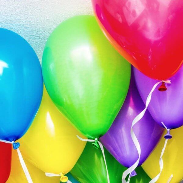 A bunch of colorful balloons against a light, textured wall. The balloons include blue, red, green, yellow, and purple colors, each with a white ribbon attached to the bottom. Sourced from top helium suppliers in Metro Manila, they are closely clustered together, creating a vibrant and festive display.