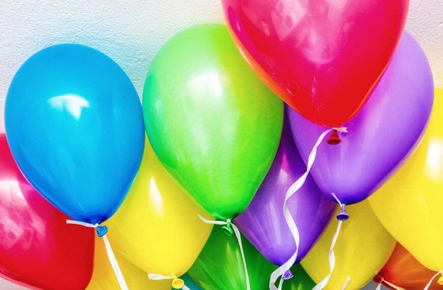 A bunch of colorful balloons against a light, textured wall. The balloons include blue, red, green, yellow, and purple colors, each with a white ribbon attached to the bottom. Sourced from top helium suppliers in Metro Manila, they are closely clustered together, creating a vibrant and festive display.