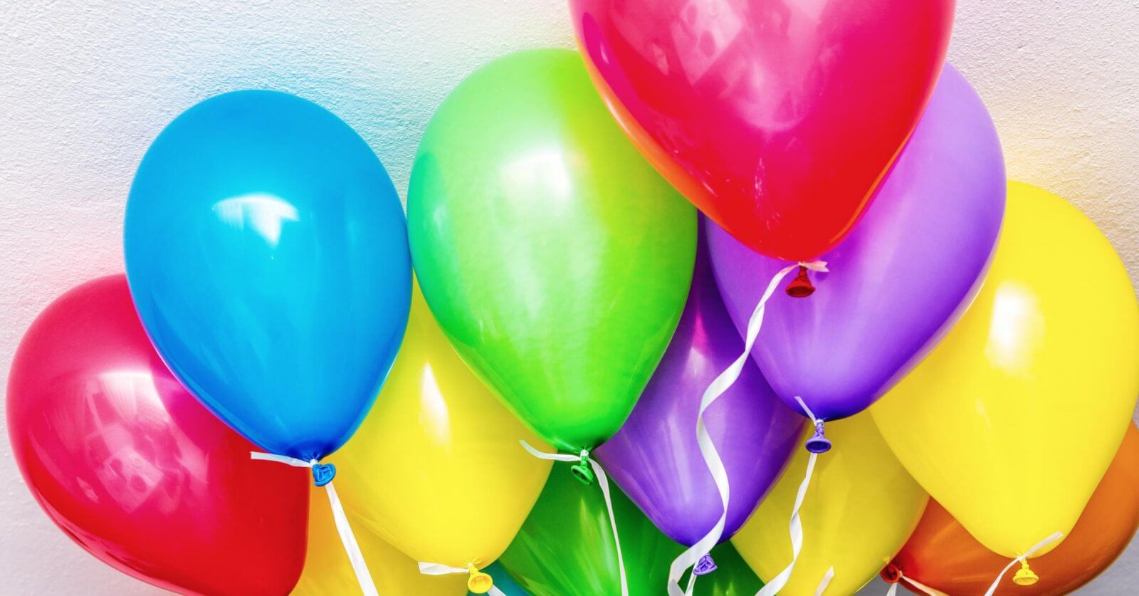A bunch of colorful balloons against a light, textured wall. The balloons include blue, red, green, yellow, and purple colors, each with a white ribbon attached to the bottom. Sourced from top helium suppliers in Metro Manila, they are closely clustered together, creating a vibrant and festive display.