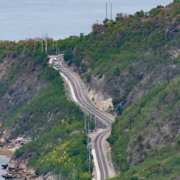 A winding road cuts through the lush, hilly terrain of Soccsksargen along the coastline. The road closely follows the contour of the land, with the sea visible on the left. Sparse greenery dots the slopes, and a few vehicles travel on this Region 12 route. The calm sea and green hills create a tranquil, scenic view.