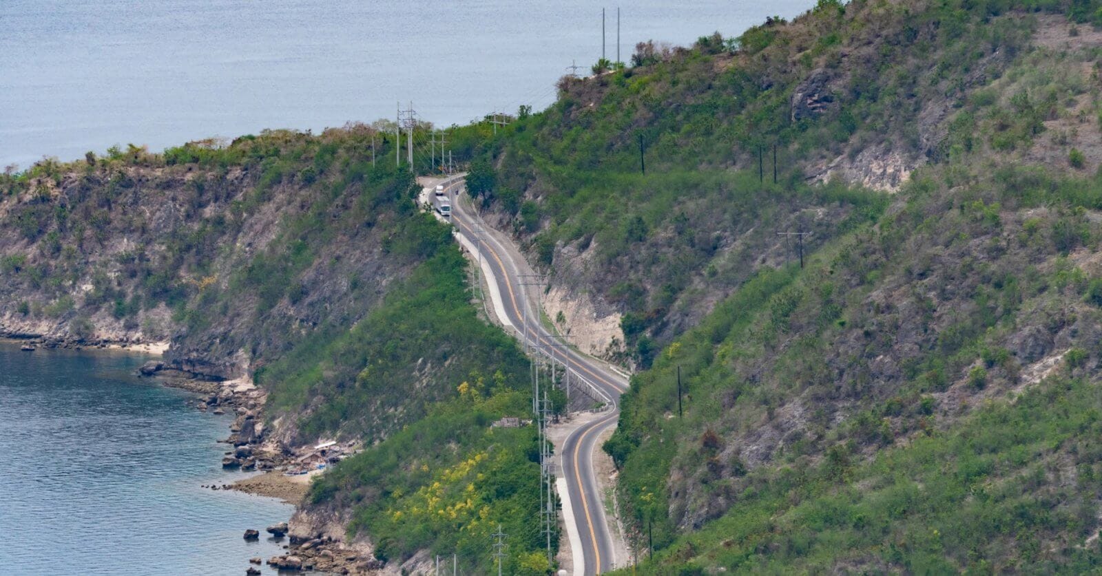 A winding road cuts through the lush, hilly terrain of Soccsksargen along the coastline. The road closely follows the contour of the land, with the sea visible on the left. Sparse greenery dots the slopes, and a few vehicles travel on this Region 12 route. The calm sea and green hills create a tranquil, scenic view.