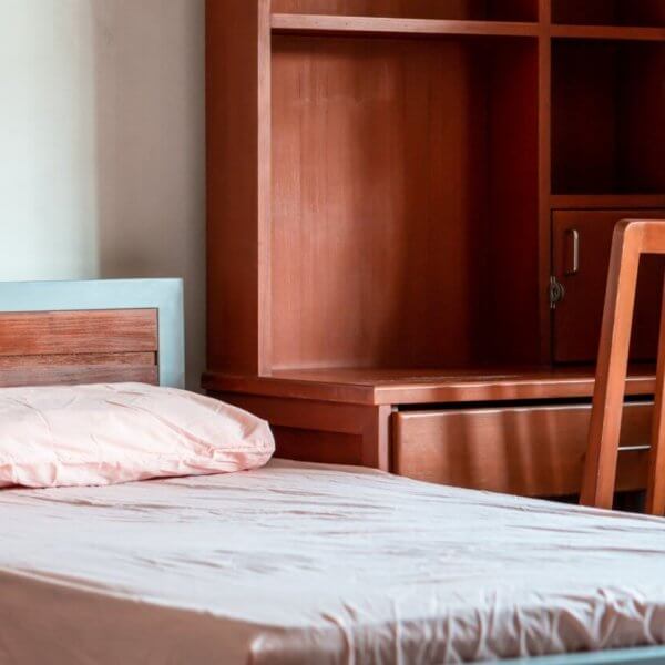 A neatly made single bed with a light pink pillow and blanket, positioned next to a wooden desk and chair set. The desk has storage compartments and a shelf unit above it. A curtain-draped window is partially visible on the right, letting in soft natural light—perfect for your stay in Metro Manila's best apartelle.