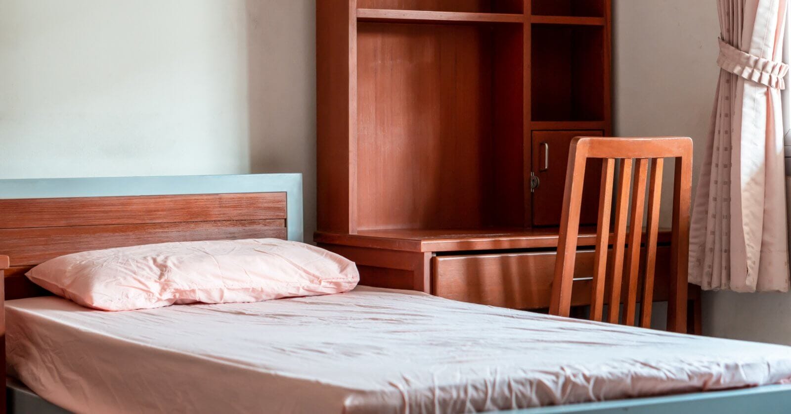 A neatly made single bed with a light pink pillow and blanket, positioned next to a wooden desk and chair set. The desk has storage compartments and a shelf unit above it. A curtain-draped window is partially visible on the right, letting in soft natural light—perfect for your stay in Metro Manila's best apartelle.