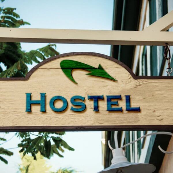 A wooden hostel sign hangs outside a building, featuring the word "HOSTEL" in blue capital letters. Above the text is a green arrow. Two flower-like designs are positioned on either side of the sign. The background includes part of a building's exterior and some tree foliage, representing one of the best hostels in Metro Manila among 49 hostels.