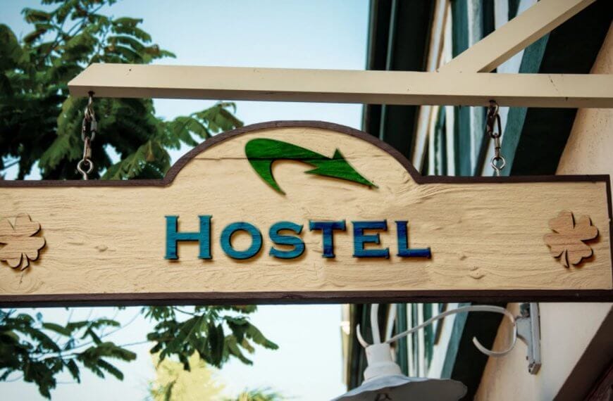 A wooden hostel sign hangs outside a building, featuring the word "HOSTEL" in blue capital letters. Above the text is a green arrow. Two flower-like designs are positioned on either side of the sign. The background includes part of a building's exterior and some tree foliage, representing one of the best hostels in Metro Manila among 49 hostels.