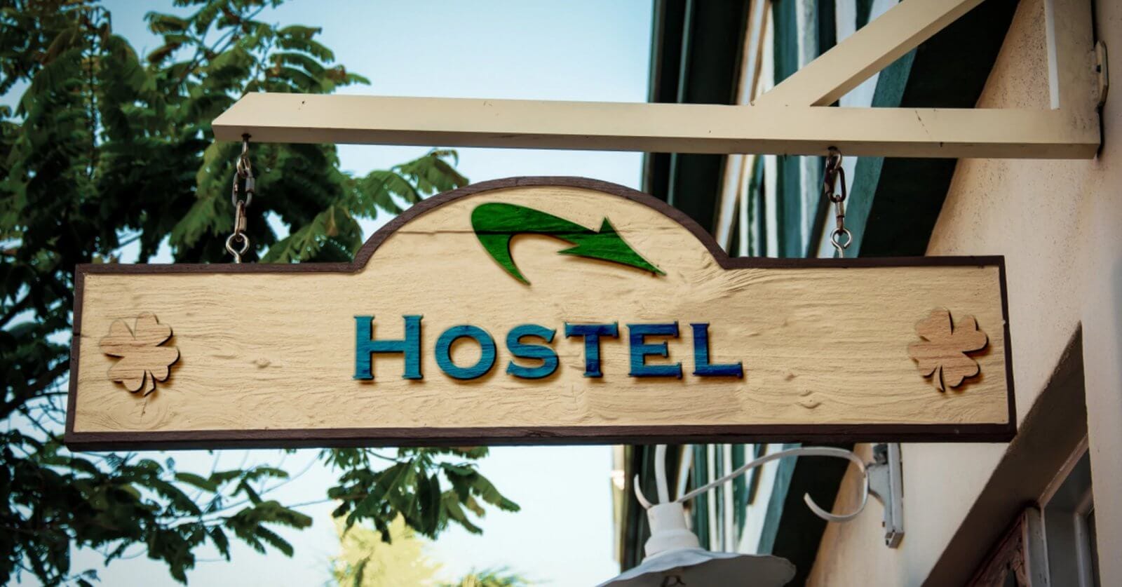A wooden hostel sign hangs outside a building, featuring the word "HOSTEL" in blue capital letters. Above the text is a green arrow. Two flower-like designs are positioned on either side of the sign. The background includes part of a building's exterior and some tree foliage, representing one of the best hostels in Metro Manila among 49 hostels.