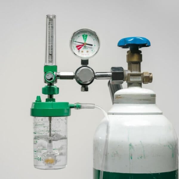 A medical oxygen tank with a pressure gauge, flow meter, and humidifier bottle attached. The tank is white with a green band near the top, and the attached components are mostly green and silver, with a blue knob. Commonly available from oxygen tank suppliers in Metro Manila, the backdrop is plain light gray.