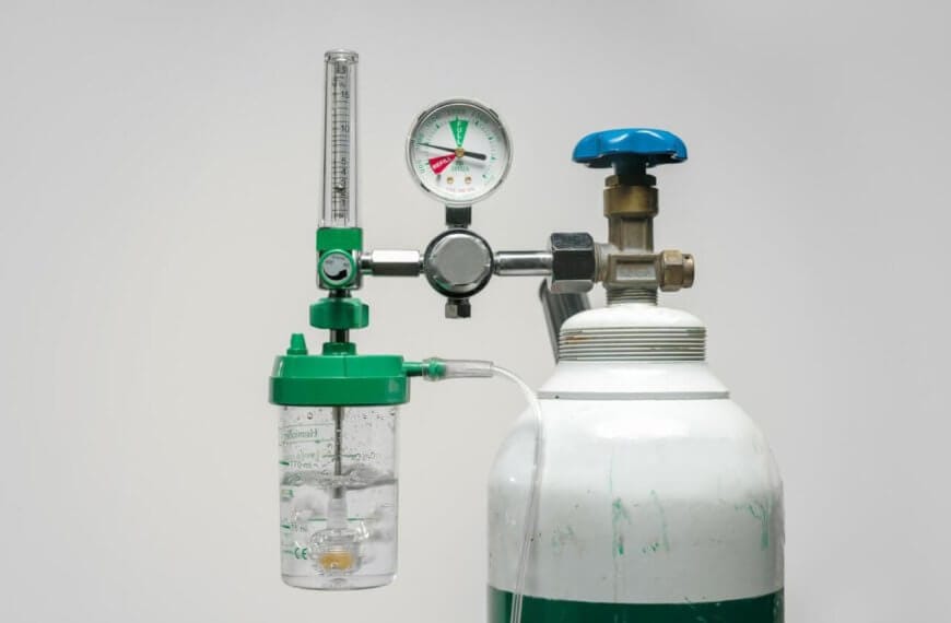 A medical oxygen tank with a pressure gauge, flow meter, and humidifier bottle attached. The tank is white with a green band near the top, and the attached components are mostly green and silver, with a blue knob. Commonly available from oxygen tank suppliers in Metro Manila, the backdrop is plain light gray.