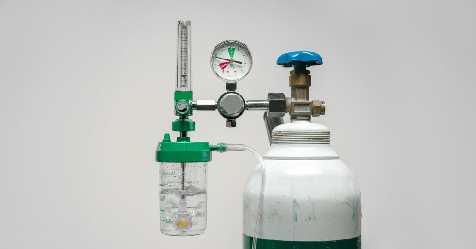 A medical oxygen tank with a pressure gauge, flow meter, and humidifier bottle attached. The tank is white with a green band near the top, and the attached components are mostly green and silver, with a blue knob. Commonly available from oxygen tank suppliers in Metro Manila, the backdrop is plain light gray.