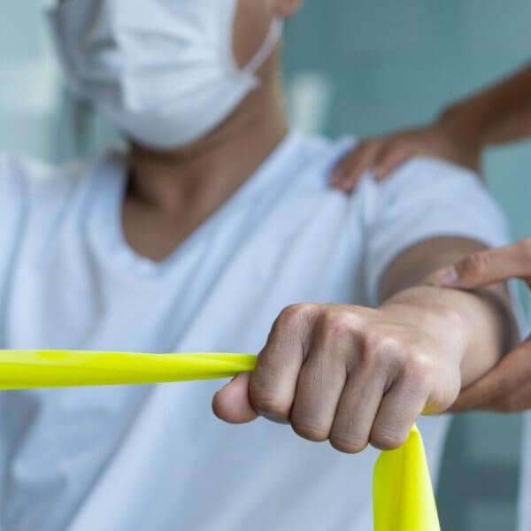 a person wearing a face mask is exercising with a yellow resistance band. Another individual, also masked, stands beside them with one hand on their back, providing support and guidance. The setting appears to be a healthcare or rehabilitation environment offering Physical Therapy Services.