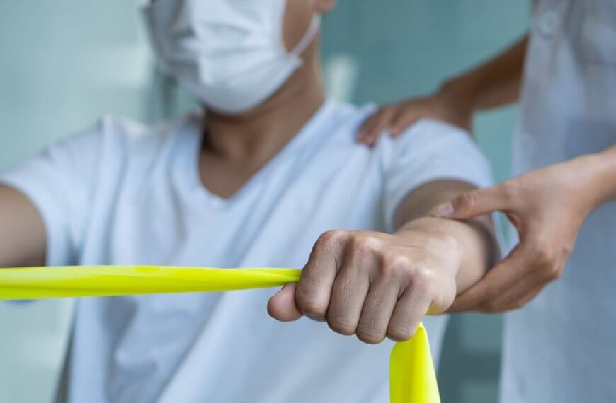 a person wearing a face mask is exercising with a yellow resistance band. Another individual, also masked, stands beside them with one hand on their back, providing support and guidance. The setting appears to be a healthcare or rehabilitation environment offering Physical Therapy Services.