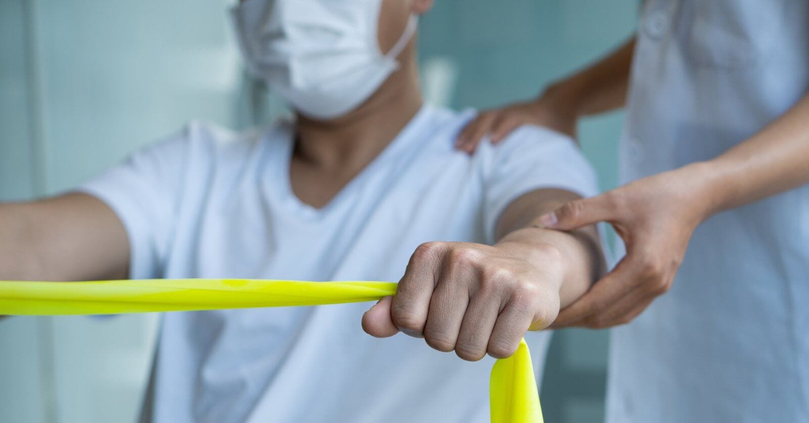 a person wearing a face mask is exercising with a yellow resistance band. Another individual, also masked, stands beside them with one hand on their back, providing support and guidance. The setting appears to be a healthcare or rehabilitation environment offering Physical Therapy Services.