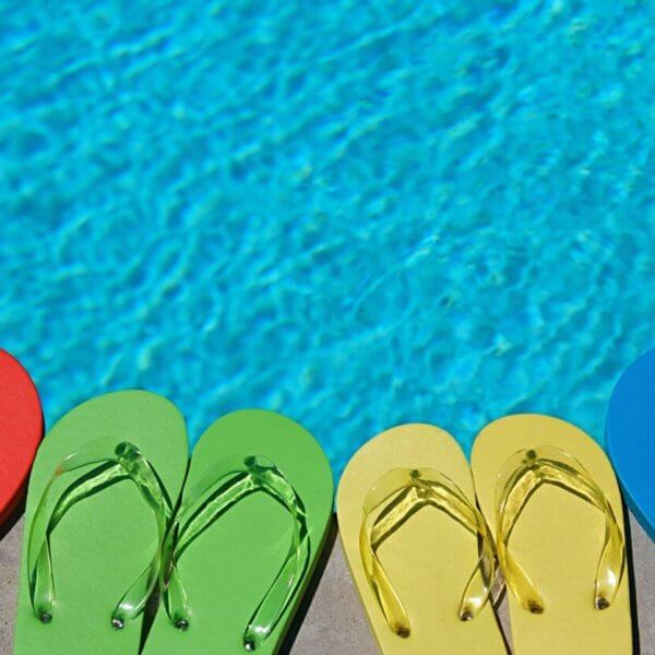 Four pairs of flip-flops lined up near a swimming pool at one of the best resorts. The flip-flops are in different colors: red, green, yellow, and blue. The clear blue pool water reflects the sunlight, evoking a bright, summery day perfect for an unforgettable escape.