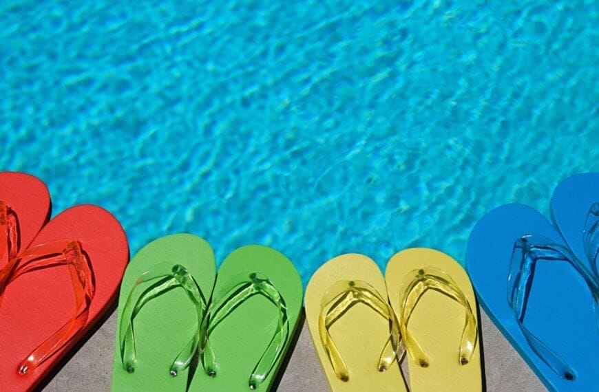 Four pairs of flip-flops lined up near a swimming pool at one of the best resorts. The flip-flops are in different colors: red, green, yellow, and blue. The clear blue pool water reflects the sunlight, evoking a bright, summery day perfect for an unforgettable escape.