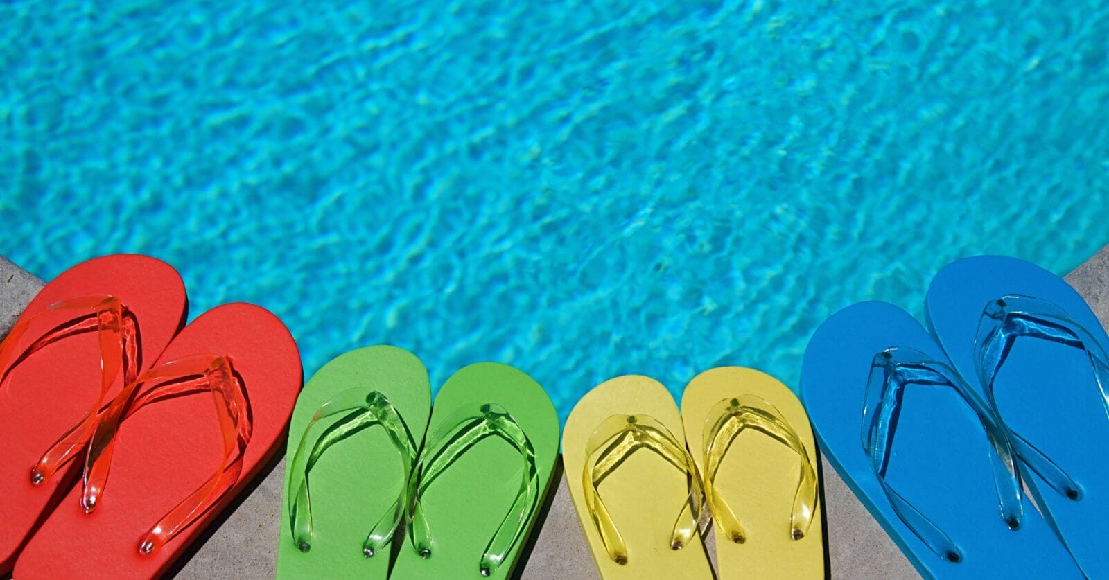 Four pairs of flip-flops lined up near a swimming pool at one of the best resorts. The flip-flops are in different colors: red, green, yellow, and blue. The clear blue pool water reflects the sunlight, evoking a bright, summery day perfect for an unforgettable escape.