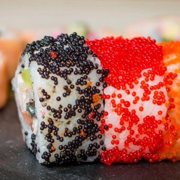 Close-up of sushi rolls from Mitsukoshi BGC decorated with black, red, and orange fish roe on a slate surface. The rolls have a visible mix of rice and seaweed, and various other ingredients inside, with out-of-focus sushi pieces seen in the background. Black chopsticks are placed to the left.
