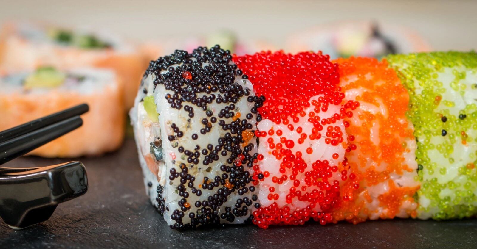 Close-up of sushi rolls from Mitsukoshi BGC decorated with black, red, and orange fish roe on a slate surface. The rolls have a visible mix of rice and seaweed, and various other ingredients inside, with out-of-focus sushi pieces seen in the background. Black chopsticks are placed to the left.