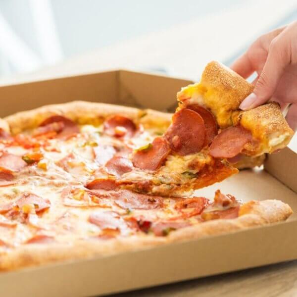 A hand is picking up a slice of pepperoni pizza from an open cardboard pizza box. The pizza has a golden-brown crust, melted cheese, and pepperoni slices. Enjoying this mouthwatering treat at one of the best restaurants in Cubao near Farmers Plaza adds to the delightful indoor setting.