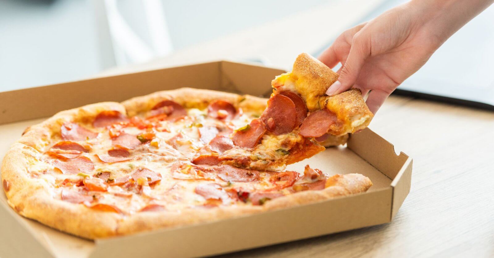 A hand is picking up a slice of pepperoni pizza from an open cardboard pizza box. The pizza has a golden-brown crust, melted cheese, and pepperoni slices. Enjoying this mouthwatering treat at one of the best restaurants in Cubao near Farmers Plaza adds to the delightful indoor setting.