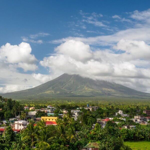 A picturesque landscape in the Bicol Region features a dormant volcano enveloped in clouds with a lush, green forest at its base. In the foreground, a small town with colorful buildings and rooftops. The sky is bright blue with scattered white clouds, creating a serene atmosphere.
