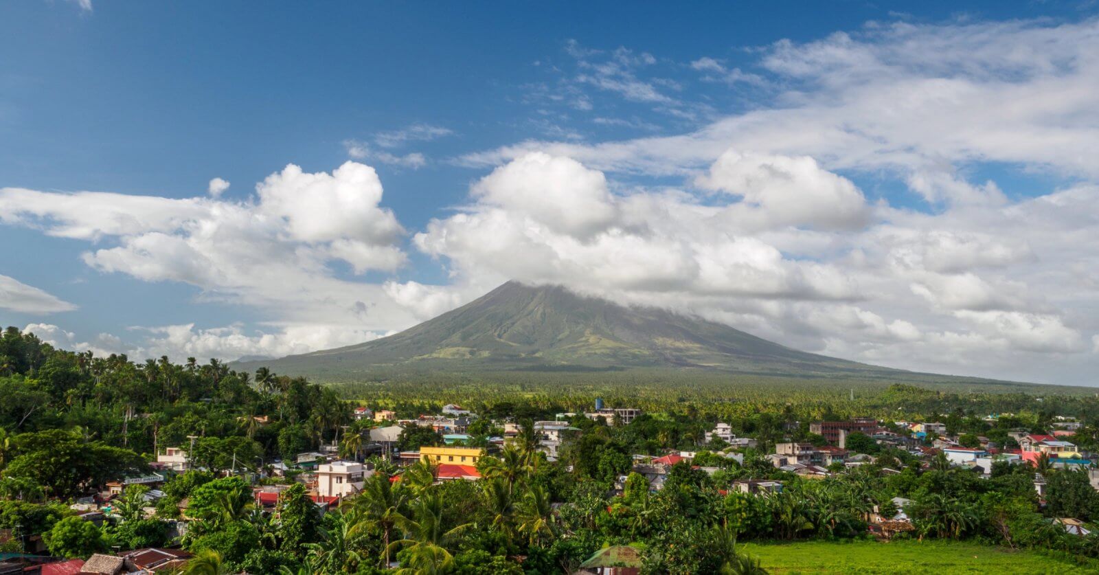 A picturesque landscape in the Bicol Region features a dormant volcano enveloped in clouds with a lush, green forest at its base. In the foreground, a small town with colorful buildings and rooftops. The sky is bright blue with scattered white clouds, creating a serene atmosphere.