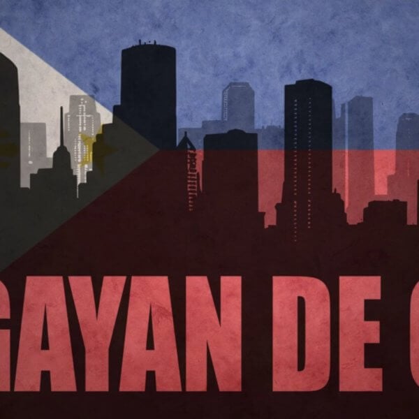 A stylized image of a city skyline with silhouettes of tall buildings. The background features the Philippine flag, blending into the skyline with its blue, red, white, and yellow colors and details. "CAGAYAN DE ORO," renowned for its tourist attractions, is prominently written in bold red letters across the bottom.