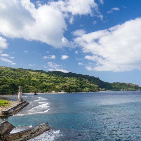 A scenic coastal landscape in Cagayan Valley with clear blue waters meets a green, hilly shoreline under a partly cloudy sky. A small lighthouse and ruins of a pier are visible to the left, while lush, verdant hills extend into the distance, providing a striking contrast to the azure sea and sky.