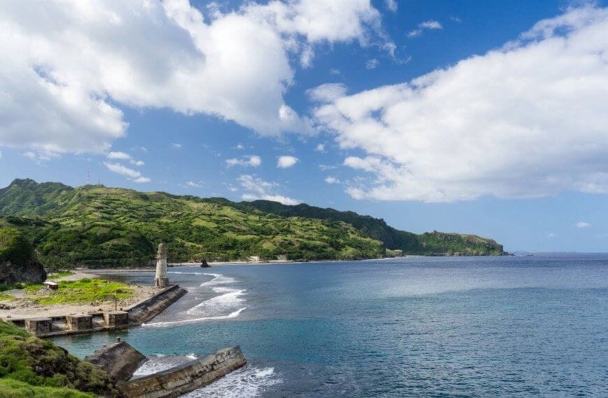 A scenic coastal landscape in Cagayan Valley with clear blue waters meets a green, hilly shoreline under a partly cloudy sky. A small lighthouse and ruins of a pier are visible to the left, while lush, verdant hills extend into the distance, providing a striking contrast to the azure sea and sky.