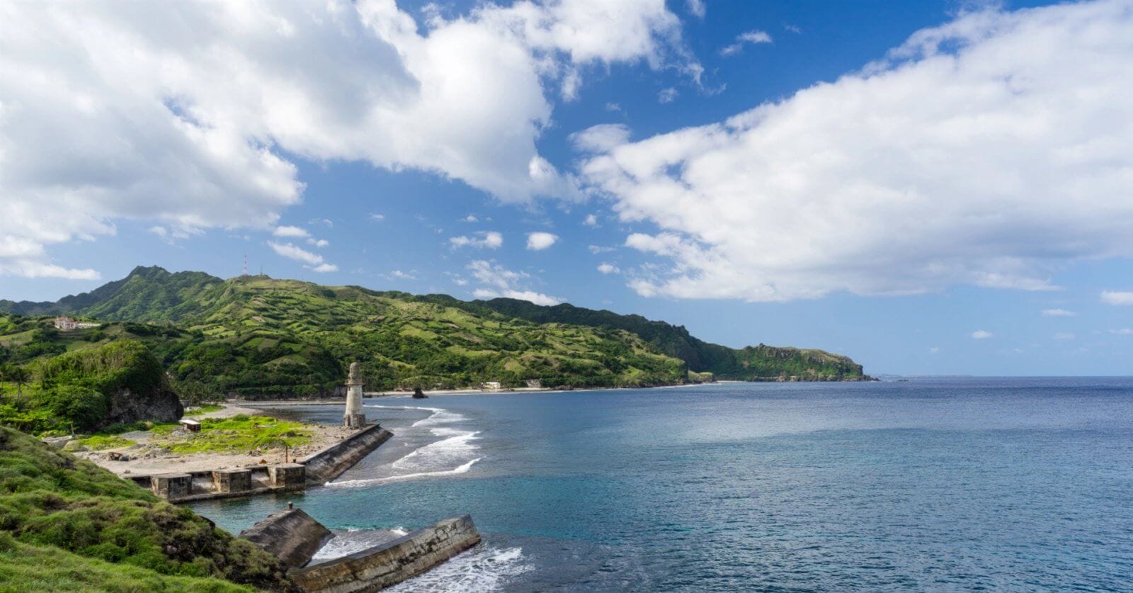 A scenic coastal landscape in Cagayan Valley with clear blue waters meets a green, hilly shoreline under a partly cloudy sky. A small lighthouse and ruins of a pier are visible to the left, while lush, verdant hills extend into the distance, providing a striking contrast to the azure sea and sky.
