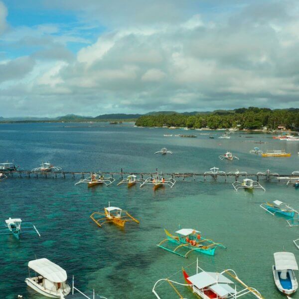 The image captures a coastal scene in the Caraga Region with numerous colorful outriggers and small boats docked or moored in clear, calm waters. The backdrop includes lush green trees, a cloudy blue sky, and distant landforms, creating a serene and picturesque maritime environment.