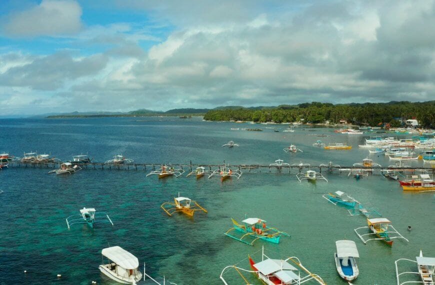 The image captures a coastal scene in the Caraga Region with numerous colorful outriggers and small boats docked or moored in clear, calm waters. The backdrop includes lush green trees, a cloudy blue sky, and distant landforms, creating a serene and picturesque maritime environment.