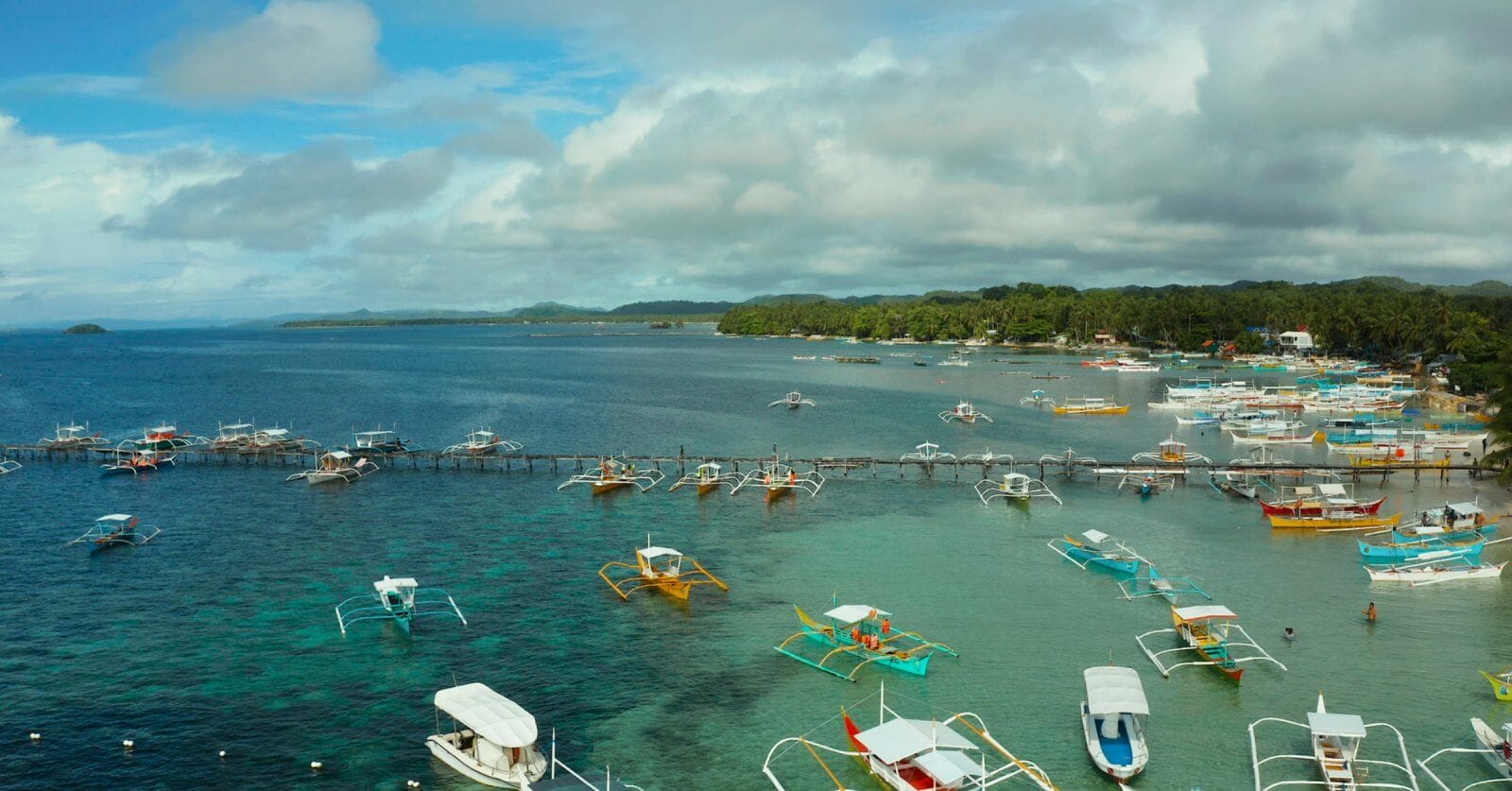 The image captures a coastal scene in the Caraga Region with numerous colorful outriggers and small boats docked or moored in clear, calm waters. The backdrop includes lush green trees, a cloudy blue sky, and distant landforms, creating a serene and picturesque maritime environment.