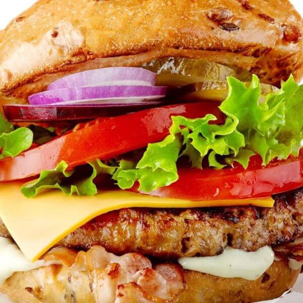 A colorful, juicy cheeseburger from one of the best restaurants in Imus Cavite is shown stacked high with layers of fresh, crisp lettuce, sliced red tomatoes, melted cheese, savory beef patty, crispy bacon, and sliced red onions. The bun is lightly toasted and speckled with sesame seeds. The burger is set against a white background.