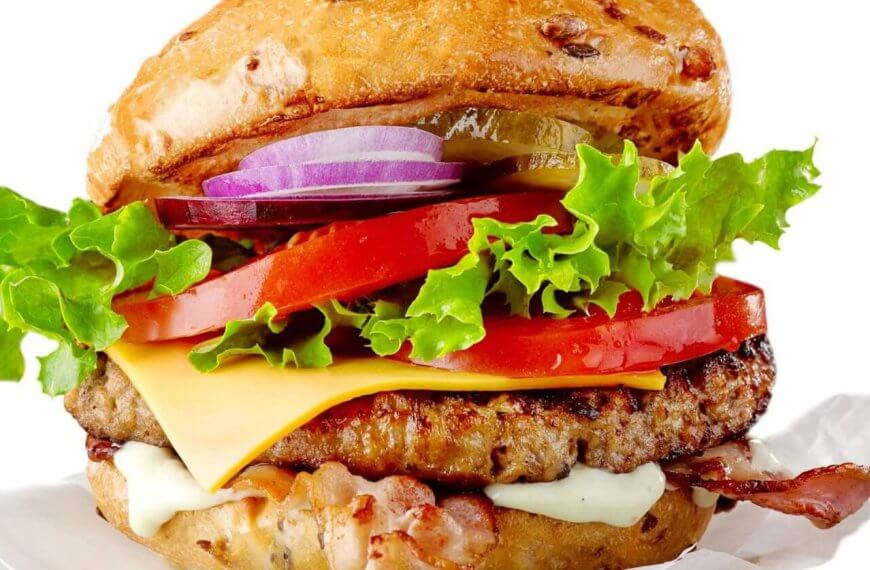 A colorful, juicy cheeseburger from one of the best restaurants in Imus Cavite is shown stacked high with layers of fresh, crisp lettuce, sliced red tomatoes, melted cheese, savory beef patty, crispy bacon, and sliced red onions. The bun is lightly toasted and speckled with sesame seeds. The burger is set against a white background.