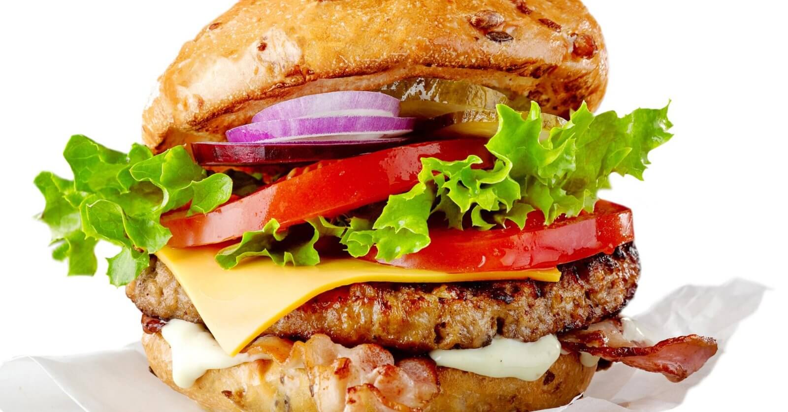 A colorful, juicy cheeseburger from one of the best restaurants in Imus Cavite is shown stacked high with layers of fresh, crisp lettuce, sliced red tomatoes, melted cheese, savory beef patty, crispy bacon, and sliced red onions. The bun is lightly toasted and speckled with sesame seeds. The burger is set against a white background.