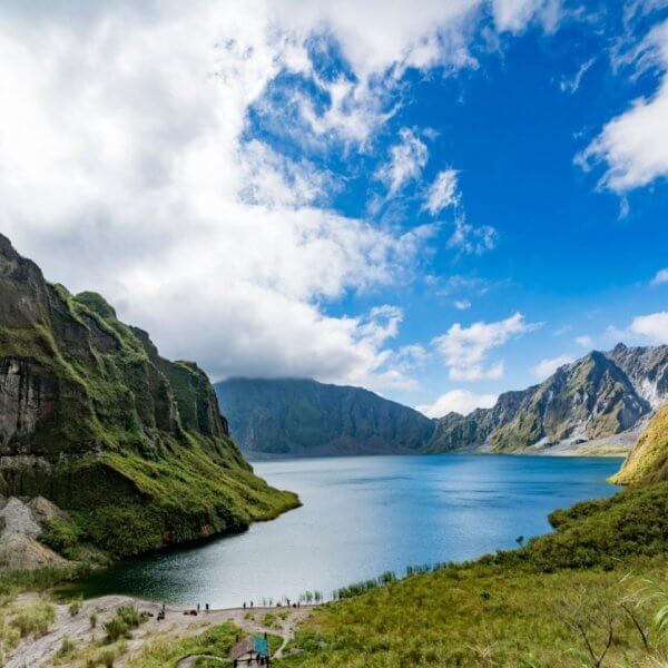 A pristine lake is nestled between towering, green-covered mountains in Region 3 under a partly cloudy sky. The vibrant blue water reflects the surrounding cliffs and the azure of the sky. Lush vegetation covers the mountainsides, and a few people are visible on a path near the lake.